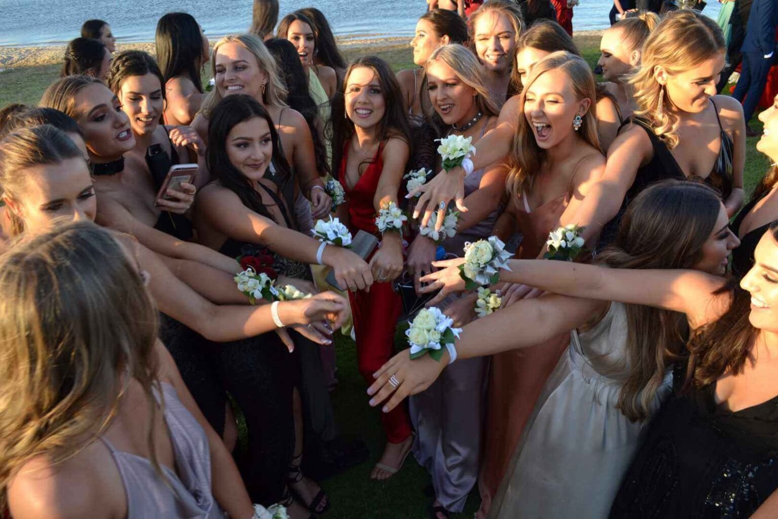 A group of excited women in formal dresses gathered in a circle, extending their hands adorned with corsages, celebrating together.