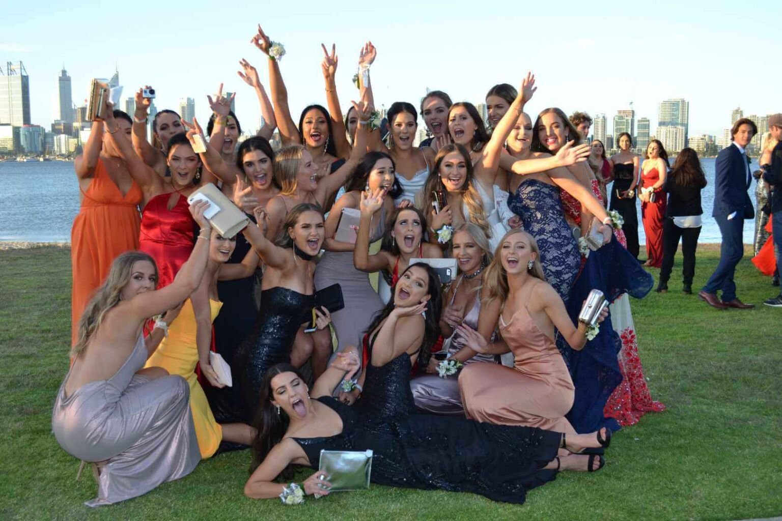 A large group of young women in colorful gowns posing joyfully for a photo on a grassy area.