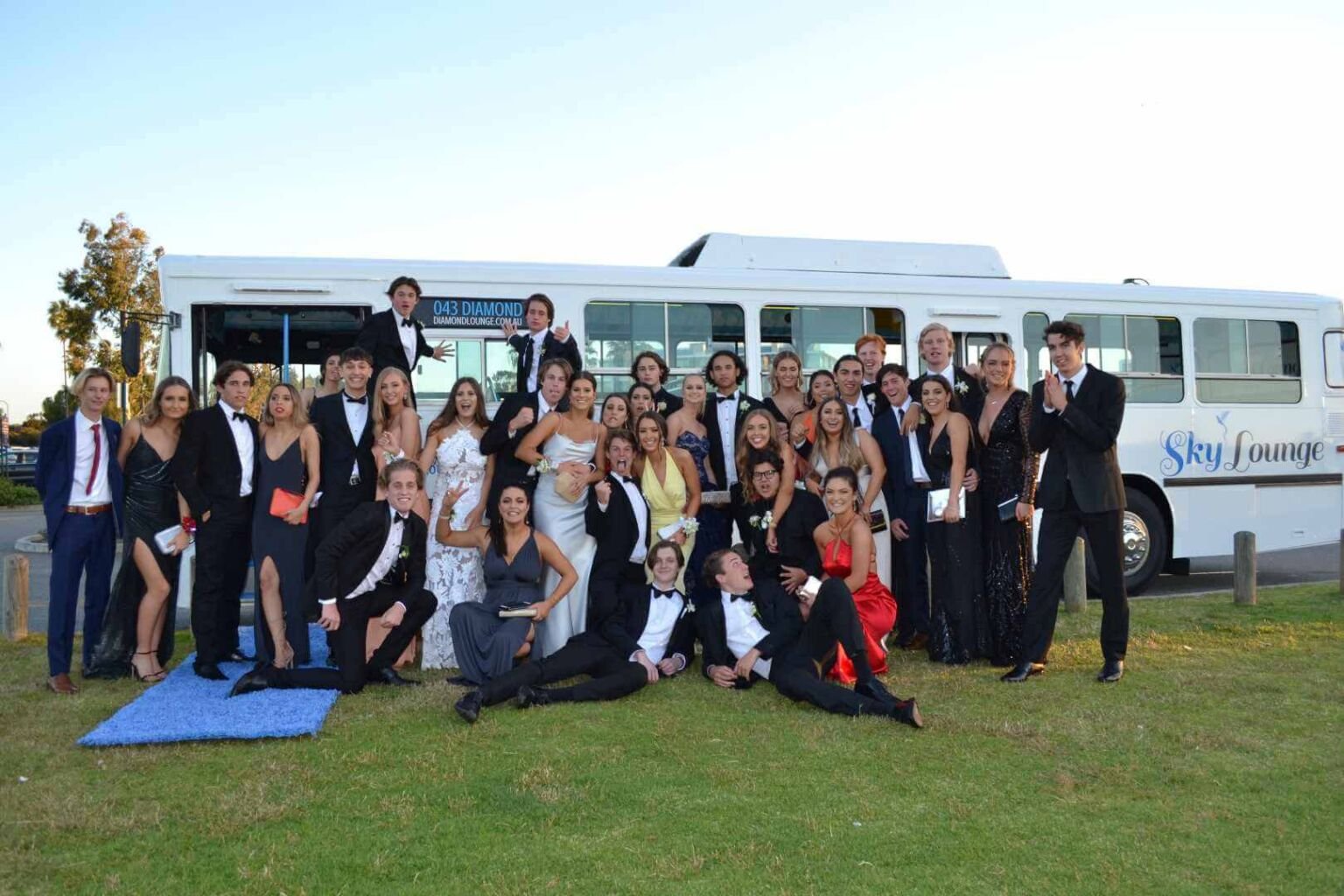 A group of young people ready for their school ball, dressed in formal attire, posing in front of a party bus labeled Sky Lounge.
