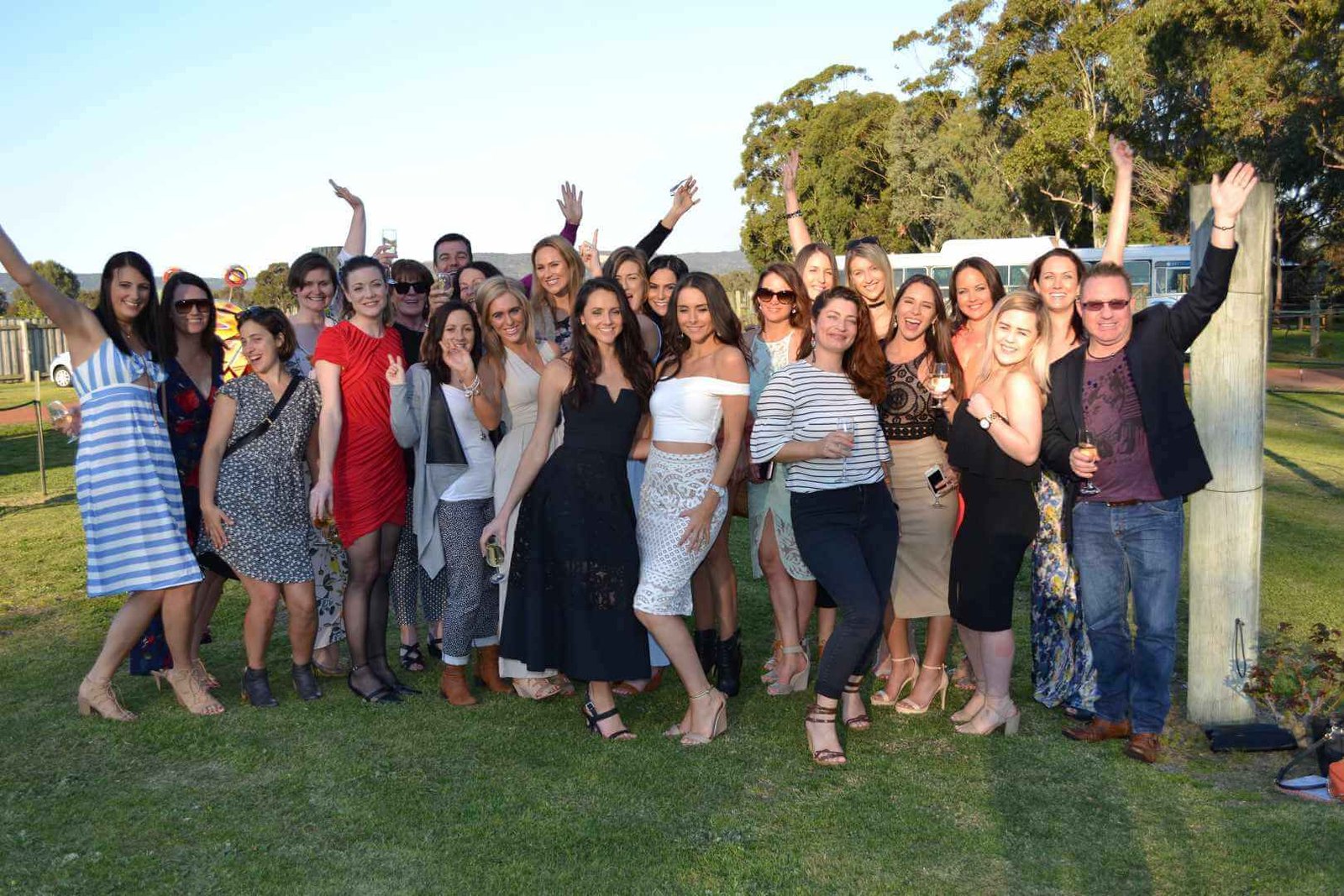 A large group of people attending a wine tasting event, stand closely on a lawn, smiling and waving.